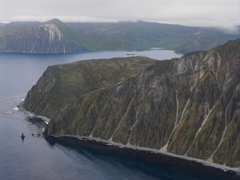 Aerial view of the rugged coastline and steep mountains of Unalaska, featuring dark rocky cliffs, deep blue waters, and a cloudy sky in Unalaska, Alaska, United States.