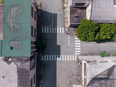 Aerial view of a street intersection with marked crosswalks, adjacent buildings with green and grey roofs, and shadows cast across the asphalt, Casale Monferrato, Piedmont, Italy.