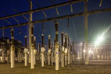 An industrial high voltage electrical substation illuminated by bright floodlights with ceramic insulators, concrete pillars, and power lines at night in Brazil. © Jose Luis Stephens