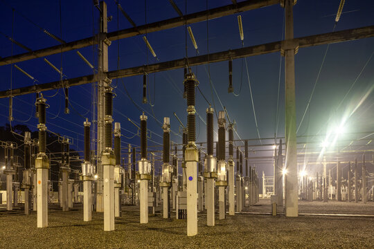 An industrial high voltage electrical substation illuminated by bright floodlights with ceramic insulators, concrete pillars, and power lines at night in Brazil.