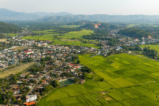 Aerial view of Lak Lake surrounded by lush green rice paddies, small village houses, and distant mountains in Lien Son Lak, Dak Lak, Vietnam.