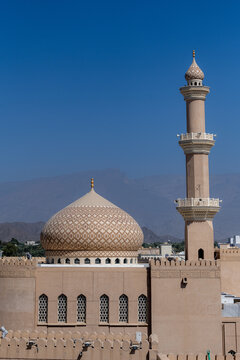 View of a mosque with an intricate patterned dome and a tall minaret set against a clear blue sky and distant mountains in Muscat, Muscat Governorate, Oman.