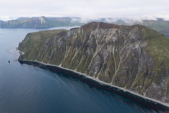 Aerial view of the rugged coastal cliffs and volcanic terrain of Unalaska, featuring steep slopes, rocky textures, and the dark waters of the Bering Sea in Unalaska, Alaska, United States.