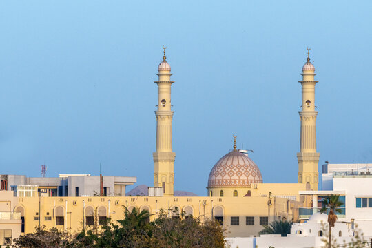 View of a mosque with two tall minarets and a patterned dome rising above urban buildings in Muscat, Muscat Governorate, Oman.