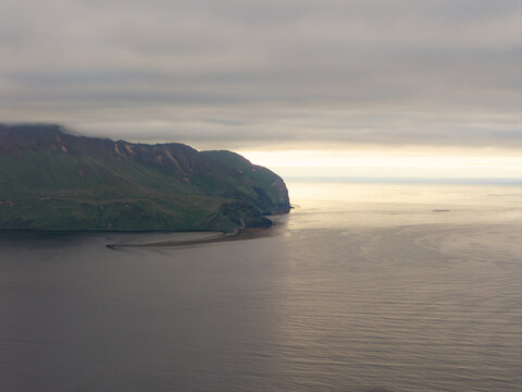 Aerial view of the rugged coastline and green hills of Unalaska under a cloudy sky at sunset in Unalaska, Alaska, United States.