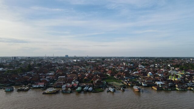 Aerial view of the Jakabaring riverside settlement with wooden houses, boats moored along the Musi River, and dense urban rooftops in Kecamatan Seberang Ulu I, Sumatera Selatan, Indonesia.
