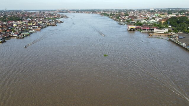 Aerial view of the Musi River with the Ampera Bridge in the distance, surrounded by dense urban housing and boats in Kecamatan Seberang Ulu I, Sumatera Selatan, Indonesia.