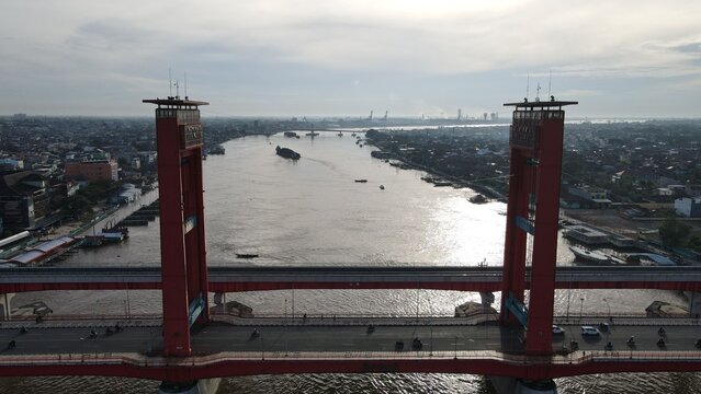 Aerial view of the iconic red Ampera Bridge spanning the Musi River with traffic flowing across and boats navigating the water in Palembang, Sumatera Selatan, Indonesia.