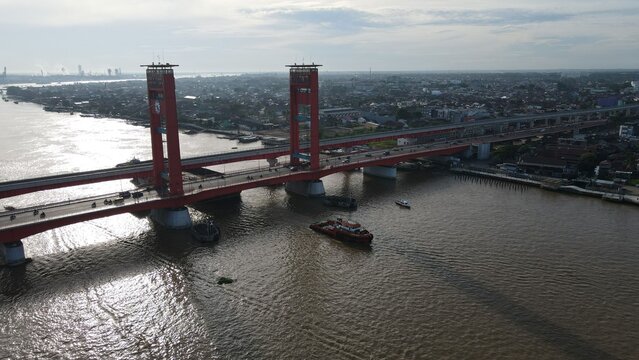 Aerial view of the red Ampera Bridge spanning the Musi River in Palembang, with urban surroundings and boats in Kecamatan Seberang Ulu I, Sumatera Selatan, Indonesia.