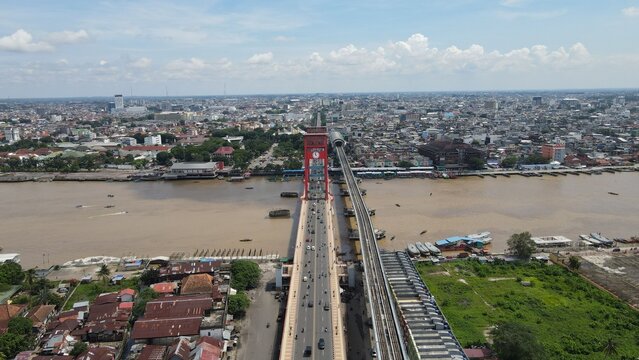Aerial view of the iconic red Ampera Bridge spanning the Musi River with urban cityscape, traffic, and railway tracks in Kecamatan Seberang Ulu I, Sumatera Selatan, Indonesia.