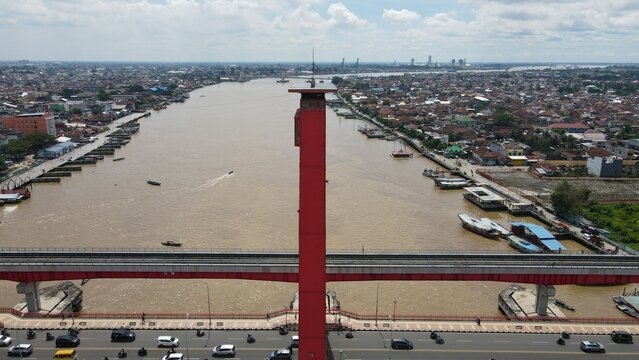 Aerial view of the red Ampera Bridge spanning the Musi River, with urban development, boats on the water, and traffic in Kecamatan Seberang Ulu I, Sumatera Selatan, Indonesia.