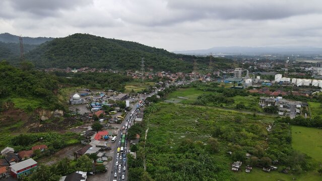 Aerial view of the Merak area featuring a busy road with traffic, lush green hills, industrial facilities, and residential buildings in Pulomerak, Banten, Indonesia.