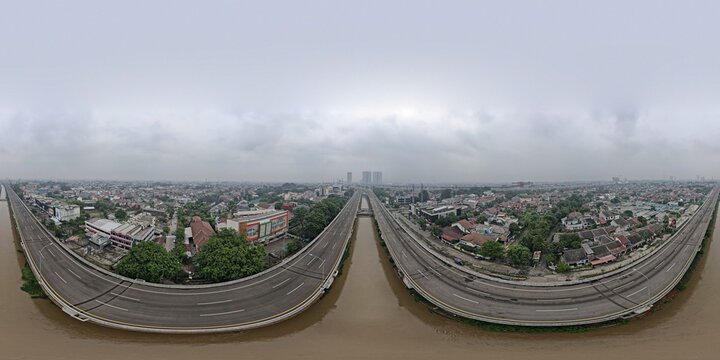 Aerial view of the elevated highway stretching over a river, surrounded by dense urban residential areas and lush greenery under a cloudy sky in Bekasi Barat, West Java, Indonesia.