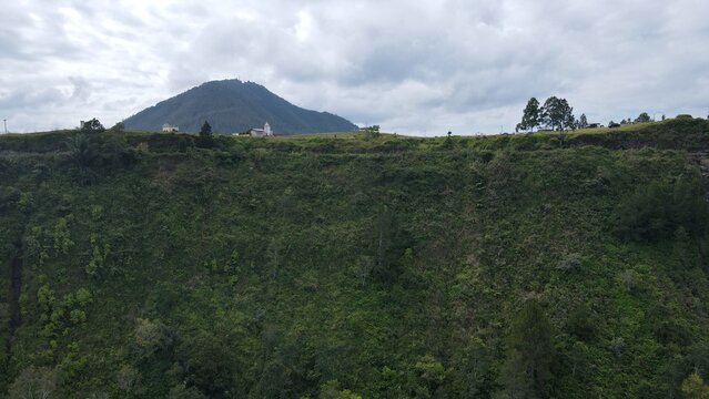 Aerial view of the Tara Bunga area featuring a small white church on a grassy ridge overlooking a steep, lush green hillside under a cloudy sky in Tampahan, North Sumatra, Indonesia.