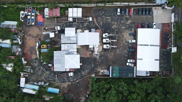 Aerial view of the Bambu Apus construction site with heavy machinery, parked trucks, and warehouse facilities on muddy ground in Kecamatan Cipayung, Daerah Khusus Ibukota, Indonesia.