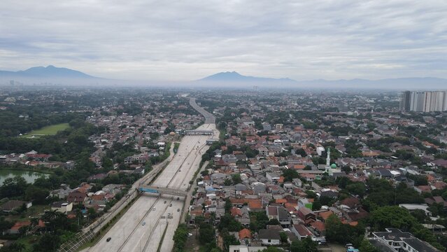 Aerial view of Jalan Tol Depok - Antasari highway cutting through the urban landscape with residential rooftops and distant mountains, Kecamatan Cilandak, Daerah Khusus Ibukota, Indonesia.