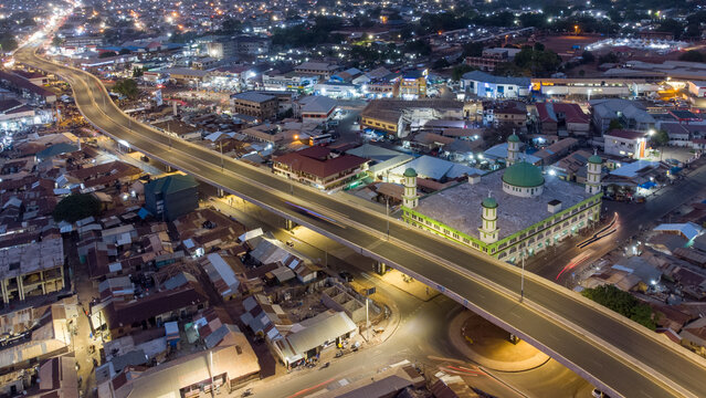 Aerial view of the illuminated Tamale Central Mosque situated next to an elevated highway with city lights and traffic streaks in the urban landscape of Tamale, Northern Region, Ghana.