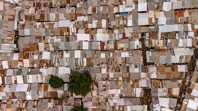 Aerial view of a dense urban neighborhood featuring a patchwork of corrugated metal rooftops in various states of rust and wear, with green trees, Tamale, Northern Region, Ghana.