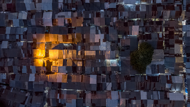 Aerial view of a dense urban neighborhood with corrugated metal rooftops and a single tree, illuminated by warm artificial light at night in Tamale, Northern Region, Ghana.