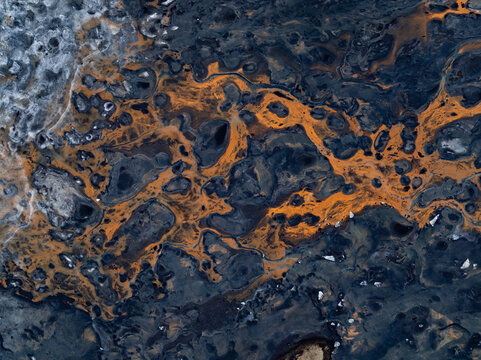Aerial view of a volcanic landscape featuring intricate patterns of dark basaltic rock and vibrant orange mineral deposits in Iceland.
