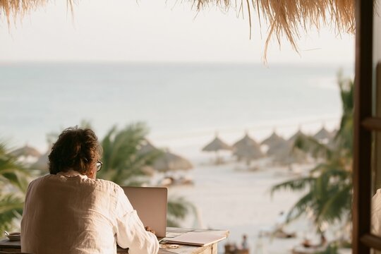 Man working on a laptop at a wooden table overlooking a tropical beach with palm trees and ocean view for a remote work and digital nomad lifestyle