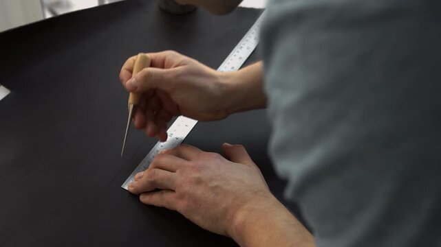 Craftsman scoring black cardboard with a metal ruler and awl for a project. Detailed artisan work in a workshop creating a box
