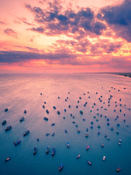Aerial view of numerous small fishing boats scattered across the calm blue sea under a vibrant pink and orange sunset sky in Vietnam.