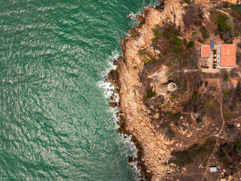 Aerial view of a lighthouse situated on a rugged rocky coastline with turquoise sea waves crashing against the cliffs and a small building with a red roof nearby in Vietnam.