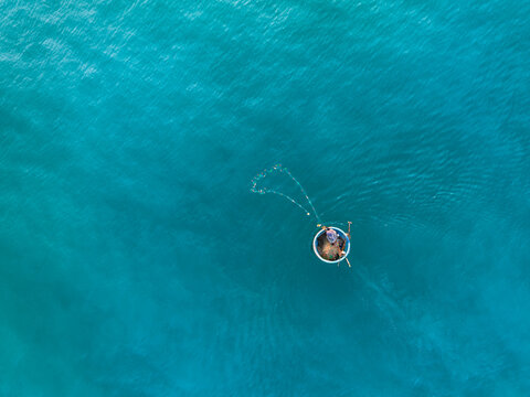Aerial view of a fisherman casting a net from a traditional round basket boat on the turquoise waters of Vietnam.