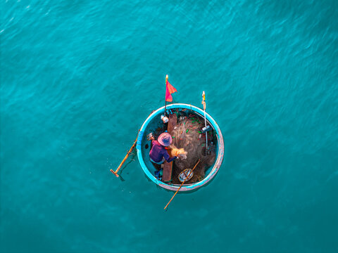 Aerial view of a fisherman working in a traditional round basket boat with fishing nets and a red flag on turquoise water in Vietnam.