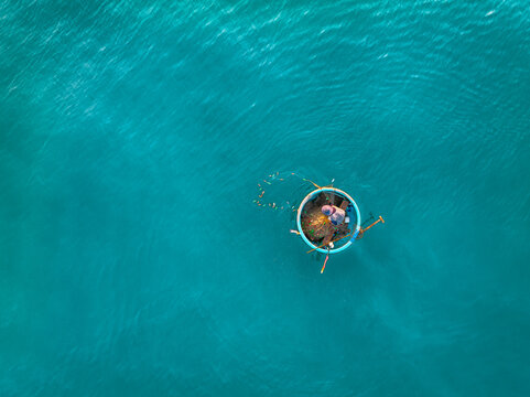 Aerial view of a fisherman in a traditional round basket boat floating on clear turquoise sea water in Vietnam.