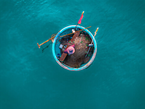 Aerial view of a traditional round basket boat with a fisherman sorting fishing nets in the turquoise waters of Vietnam.
