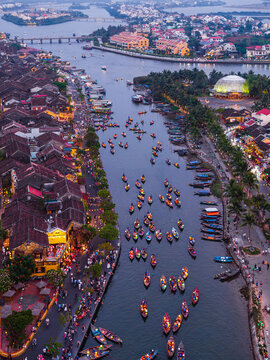 Aerial view of the Thu Bon River featuring numerous small wooden boats, illuminated historic buildings, and bustling waterfront streets at dusk in Hoi An, Vietnam.