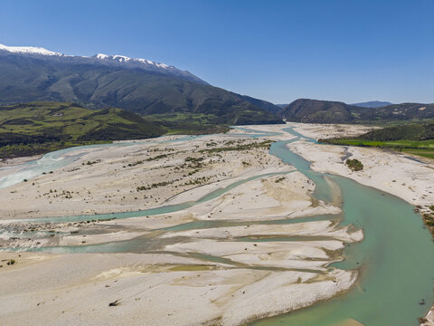 Aerial view of Vjosa Wild River National Park with its characteristic braided river channels, gravel bars, and snow-capped mountains under a clear blue sky in Permet, Gjirokaster County, Albania.