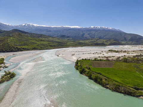 Aerial view of Vjosa Wild River National Park with turquoise river, gravel banks, green fields, and snow-capped mountains under a clear blue sky in Permet, Gjirokaster County, Albania.