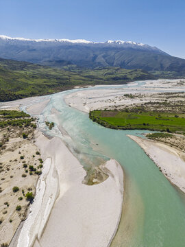 Aerial view of Vjosa Wild River National Park with turquoise river waters, expansive gravel banks, and snow-capped mountains in the background near Permet, Gjirokaster County, Albania.