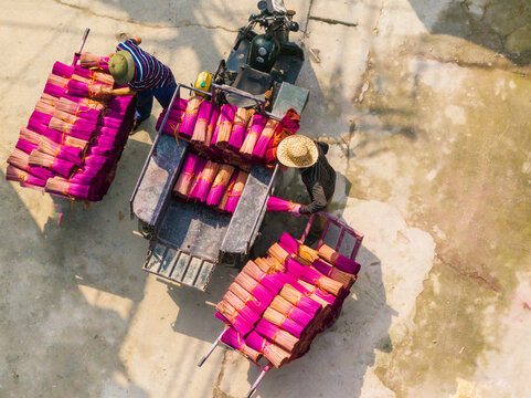 Aerial view of workers loading bundles of bright pink incense sticks onto a motorcycle cart on a concrete surface in Vietnam.