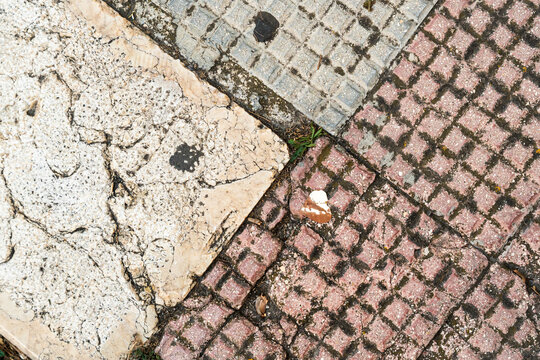 Cracked concrete slab beside textured cobblestone pavement with red and gray square pattern, weathered urban sidewalk surface showing aged deterioration and repair