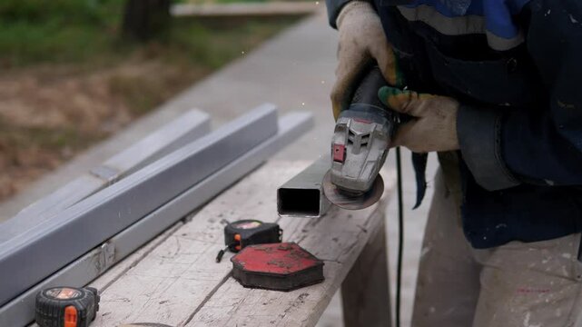 Close view shows gloved worker using handheld grinder on square metal tubing outdoors, with bright sparks scattering from contact point beside stacked steel and weathered wood surface