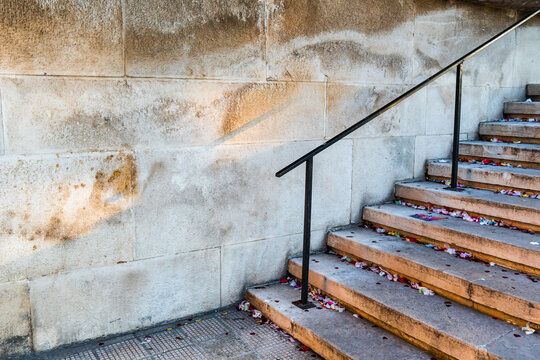 Concrete stairs with metal handrail alongside weathered stone wall with stains and rust marks, scattered colorful confetti on steps, urban architecture detail