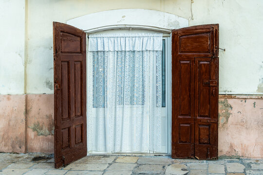 Old wooden double doors open wide on weathered building facade revealing white lace curtain behind, architecture detail of vintage colonial entrance in historic town