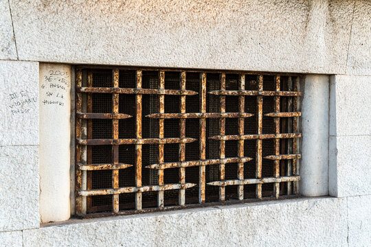 Rusty iron security grate covering basement window embedded in rough granite stone wall, with faded graffiti inscriptions on side, concept of decay and urban architecture