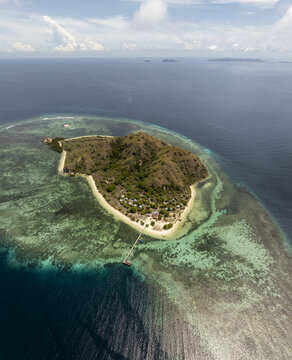 Aerial view of Kanawa Island with its lush green hills, white sandy beach, wooden jetty, and surrounding turquoise coral reefs in Kecamatan Komodo, Nusa Tenggara Timur, Indonesia.