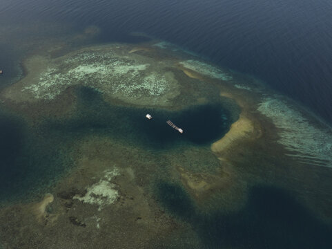 Aerial view of Kanawa Island with vibrant coral reefs, shallow turquoise waters, and deep blue sea surrounding a floating structure in Kecamatan Komodo, Nusa Tenggara Timur, Indonesia.
