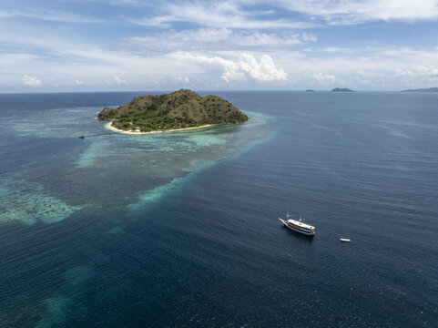 Aerial view of Kanawa Island surrounded by turquoise coral reefs and deep blue sea with a traditional wooden boat anchored nearby in Kecamatan Komodo, Nusa Tenggara Timur, Indonesia.