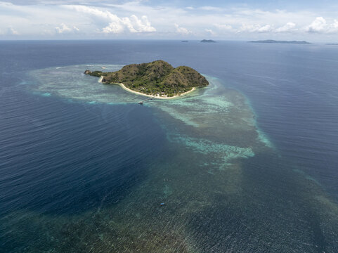 Aerial view of Kanawa Island surrounded by turquoise coral reefs and deep blue ocean waters under a partly cloudy sky in Kecamatan Komodo, Nusa Tenggara Timur, Indonesia.