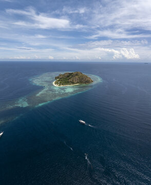 Aerial view of Kanawa Island surrounded by turquoise coral reefs and deep blue sea with boats in the distance, Kecamatan Komodo, Nusa Tenggara Timur, Indonesia.