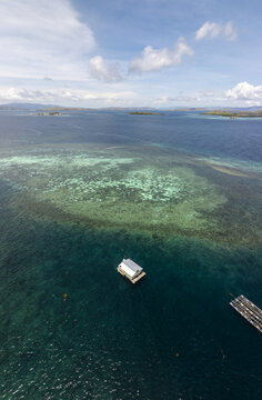 Aerial view of a small house on stilts surrounded by vibrant coral reefs and clear turquoise waters near Kanawa Island in Kecamatan Komodo, Nusa Tenggara Timur, Indonesia.