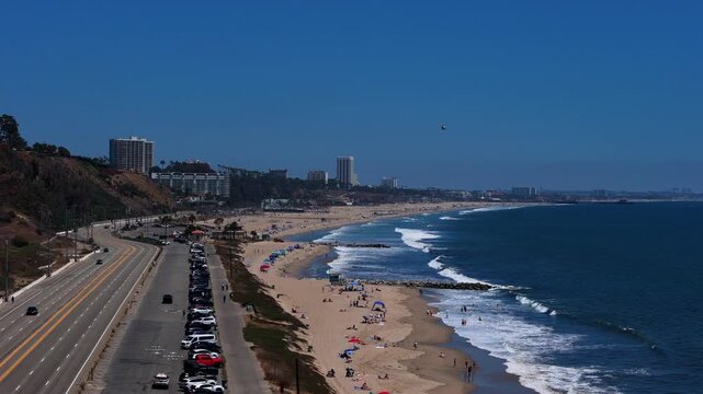 Aerial view of Will Rogers State Beach with Pacific Coast Highway, parking lot, and ocean waves under a clear blue sky in Los Angeles, California, United States.