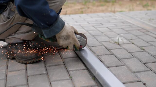 Worker crouches close to ground while grinding metal edging beside paved brick walkway, with bright sparks at contact point, muted gray tones, rough texture, sparse grass, and practical labor outdoors